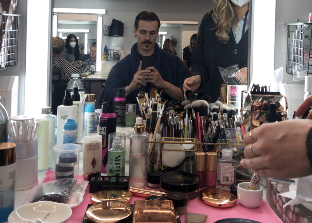 Ellis sits in a make up chair surrounded by make up
