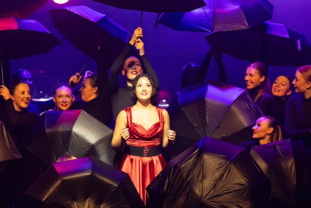Performers College student surrounded by supporting performers on stage with umbrellas.