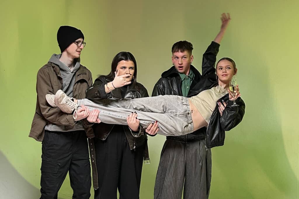 Four Performers College acting students pose in a studio, three holding a fourth horizontally aloft against a green backdrop
